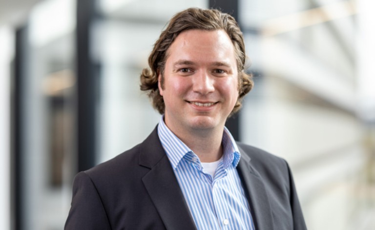 Das Porträtfoto zeigt einen jungen Mann in einer modernen Büroumgebung.
Person und Erscheinung: Der Mann blickt mit einem offenen, freundlichen Lächeln direkt in die Kamera. Er hat welliges, braunes Haar, das bis zu den Ohren reicht, und eine sympathische Ausstrahlung.
Kleidung: Er trägt ein dunkelgraues Sakko über einem hellblau-weiß fein gestreiften Business-Hemd. Das Hemd ist am Kragen offen geknöpft, was den professionellen, aber nahbaren Look unterstreicht.
Hintergrund und Komposition: Der Mann ist im Halbprofil dargestellt und leicht nach rechts versetzt positioniert. Der Hintergrund zeigt eine helle, unscharfe Büroarchitektur mit Fensterfronten und dunklen Rahmen (Bokeh-Effekt). Das Licht fällt weich von vorne links ein und leuchtet sein Gesicht gleichmäßig aus.