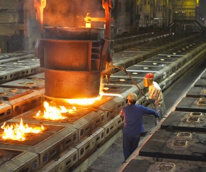 Das Foto zeigt eine beeindruckende Innenansicht einer Gießerei oder eines Stahlwerks während des Gießvorgangs von flüssigem Metall.
Zentrales Element: Eine massive, dunkle Gießpfanne hängt an einem mächtigen Deckenkran im oberen Bildzentrum. Aus der Pfanne ergießt sich ein heller, orange-gelb leuchtender Strom aus flüssigem Eisen oder Stahl in eine Reihe von Gussformen, die auf dem Boden angeordnet sind. Über der Pfanne steigen Hitze und leichter Dampf auf.
Personen und Arbeit: Zwei Stahlarbeiter in Schutzkleidung steuern den Prozess im Vordergrund.
Ein Arbeiter rechts trägt einen silbernen Hitzeschutzanzug, einen roten Schutzhelm und ein golden verspiegeltes Visier zum Schutz gegen die extreme Strahlungshitze. Er hantiert mit einer langen Stange an der Gießpfanne.
Ein zweiter Arbeiter in blauer Arbeitskleidung steht links von ihm und unterstützt den Vorgang mit einem langen Werkzeug.
Anlage und Umgebung: Die Gussformen sind in langen, parallelen Reihen auf dem Werkshallenboden aufgestellt. In einigen bereits befüllten Formen im linken Vordergrund lodern helle Flammen auf. Die Halle im Hintergrund ist weitläufig und eher dunkel, mit vereinzelten gelben Lichtern an der Decke und einer metallenen Treppenkonstruktion.
Atmosphäre: Das Bild vermittelt durch die starken Kontraste zwischen der dunklen Halle und dem intensiv leuchtenden, flüssigen Metall eine Hitze ausstrahlende und hochindustrielle Arbeitsumgebung.