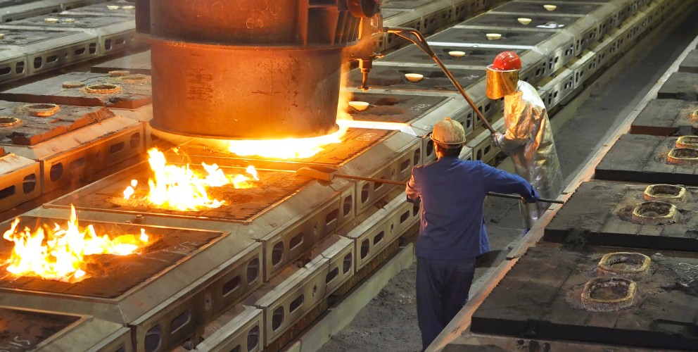 Das Foto zeigt eine beeindruckende Innenansicht einer Gießerei oder eines Stahlwerks während des Gießvorgangs von flüssigem Metall.
Zentrales Element: Eine massive, dunkle Gießpfanne hängt an einem mächtigen Deckenkran im oberen Bildzentrum. Aus der Pfanne ergießt sich ein heller, orange-gelb leuchtender Strom aus flüssigem Eisen oder Stahl in eine Reihe von Gussformen, die auf dem Boden angeordnet sind. Über der Pfanne steigen Hitze und leichter Dampf auf.
Personen und Arbeit: Zwei Stahlarbeiter in Schutzkleidung steuern den Prozess im Vordergrund.
Ein Arbeiter rechts trägt einen silbernen Hitzeschutzanzug, einen roten Schutzhelm und ein golden verspiegeltes Visier zum Schutz gegen die extreme Strahlungshitze. Er hantiert mit einer langen Stange an der Gießpfanne.
Ein zweiter Arbeiter in blauer Arbeitskleidung steht links von ihm und unterstützt den Vorgang mit einem langen Werkzeug.
Anlage und Umgebung: Die Gussformen sind in langen, parallelen Reihen auf dem Werkshallenboden aufgestellt. In einigen bereits befüllten Formen im linken Vordergrund lodern helle Flammen auf. Die Halle im Hintergrund ist weitläufig und eher dunkel, mit vereinzelten gelben Lichtern an der Decke und einer metallenen Treppenkonstruktion.
Atmosphäre: Das Bild vermittelt durch die starken Kontraste zwischen der dunklen Halle und dem intensiv leuchtenden, flüssigen Metall eine Hitze ausstrahlende und hochindustrielle Arbeitsumgebung.