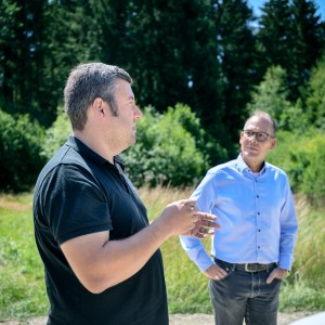 Zwei Männer im Gespräch im Freien vor einem Waldhintergrund.