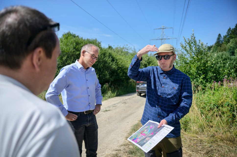 Ein Mann im schwarzen Poloshirt steht im Profil im Vordergrund und gestikuliert beim Sprechen. Im Hintergrund hört ihm ein Brillenträger im hellblauen Hemd aufmerksam zu. Sie befinden sich im Freien vor einer Kulisse aus Bäumen und Wiesen.