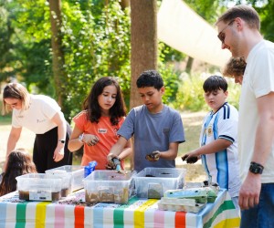 Drei Kinder befüllen an einem bunten Tisch Eierkartons mit Erde, beobachtet von zwei Erwachsenen im Freien.