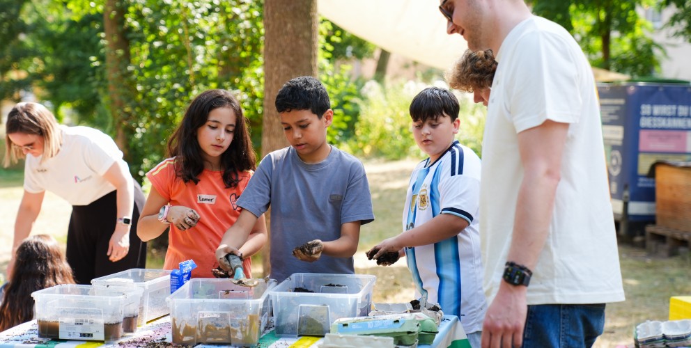 Drei Kinder befüllen an einem bunten Tisch Eierkartons mit Erde, beobachtet von zwei Erwachsenen im Freien.