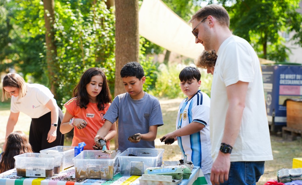 Drei Kinder befüllen an einem bunten Tisch Eierkartons mit Erde, beobachtet von zwei Erwachsenen im Freien.