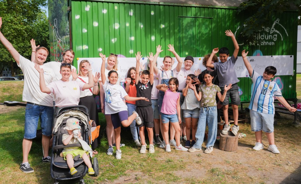 Eine fröhliche Gruppe aus Kindern und Erwachsenen posiert mit erhobenen Händen für ein Gruppenfoto vor einem grünen Bauwagen im Freien.
