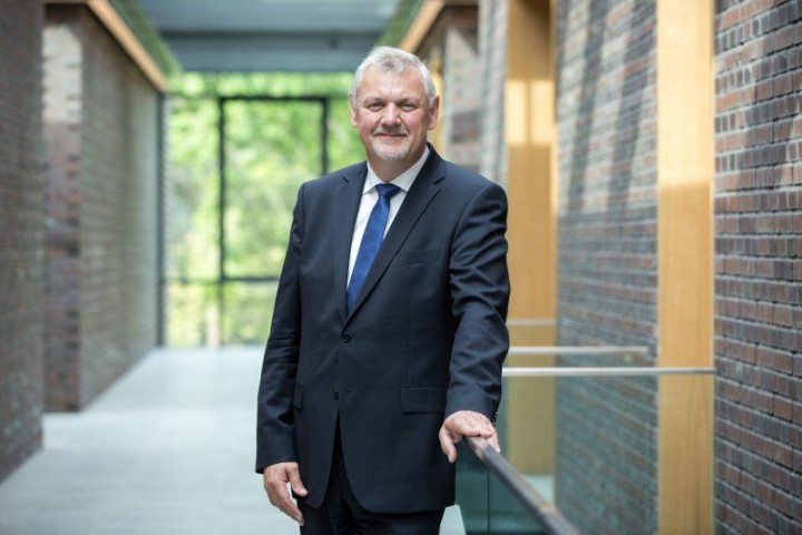 Das Bild zeigt Herrn Joachim Vanzetta. Vanzetta trägt einen dunklen Anzug und steht an ein Geländer gelehnt in einem Backsteingebäude.