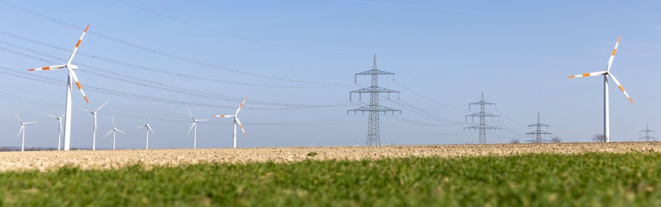 Das freundliche Foto im Querformat zeigt. Windkraftanlagen und eine Stromtrasse vor blauem Himmel. Im Vordergrund ein abgeerntetes Feld. 