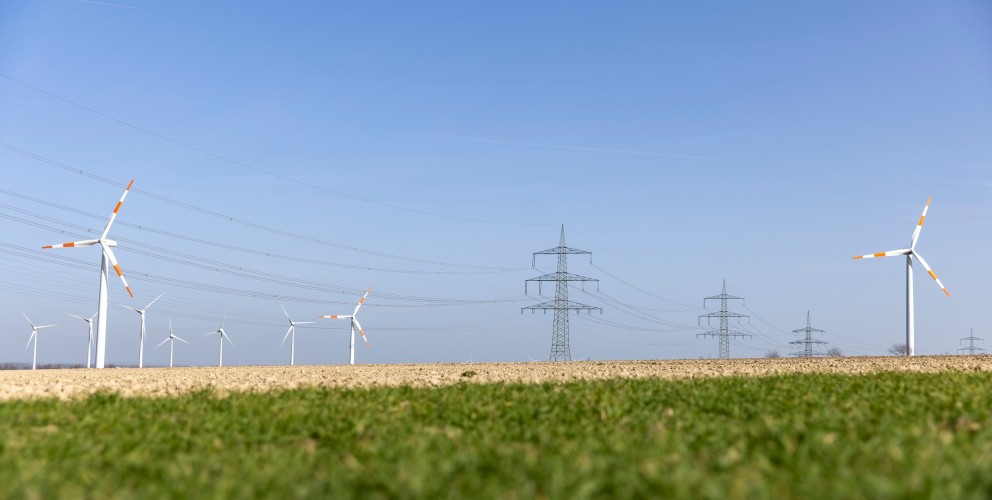Das freundliche Foto im Querformat zeigt. Windkraftanlagen und eine Stromtrasse vor blauem Himmel. Im Vordergrund ein abgeerntetes Feld. 