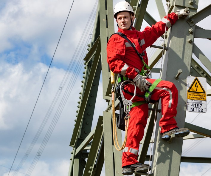 Das Bild im Querformat zeigte einen Montuer der Amprion GmbH an einem Höchstspannungsmasten. Er trägt rote Bekleidung, eine PSA und einen weißen Helm.