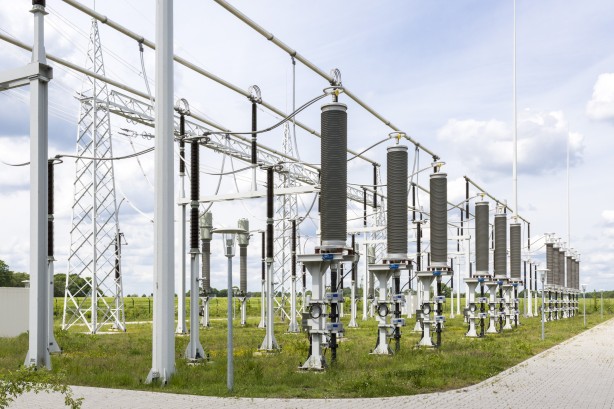 Vordergrund: Im Zentrum stehen drei große, vertikale Stützisolatoren in einer Reihe. Diese bestehen aus grauen, gerippten Zylindern, die auf stabilen, hellgrauen Metallfüßen im Gras montiert sind. An der Basis jedes Isolators befindet sich ein kleiner grauer Schaltkasten mit technischen Anschlüssen.
Mittel- und Hintergrund: Hinter der Hauptreihe sind weitere elektrische Komponenten, Isolatoren und hohe Gittermasten aus Metall zu sehen, die ein komplexes Netz aus horizontal verlaufenden Stromschienen und Leitungen tragen. Links ist eine schlichte, moderne Straßenlaterne mit einem flachen, runden Schirm platziert.
Umgebung: Die Anlage befindet sich auf einer eingezäunten Grasfläche mit grüner Vegetation und einigen gelben Wildblumen. Im Hintergrund erstreckt sich eine flache, grüne Landschaft mit Bäumen unter einem weiten, hellen Himmel.
