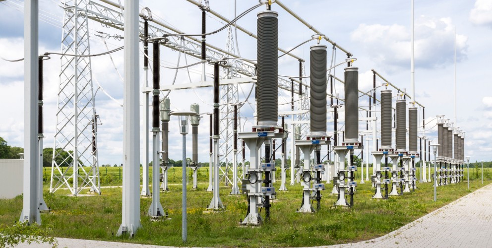 Vordergrund: Im Zentrum stehen drei große, vertikale Stützisolatoren in einer Reihe. Diese bestehen aus grauen, gerippten Zylindern, die auf stabilen, hellgrauen Metallfüßen im Gras montiert sind. An der Basis jedes Isolators befindet sich ein kleiner grauer Schaltkasten mit technischen Anschlüssen.
Mittel- und Hintergrund: Hinter der Hauptreihe sind weitere elektrische Komponenten, Isolatoren und hohe Gittermasten aus Metall zu sehen, die ein komplexes Netz aus horizontal verlaufenden Stromschienen und Leitungen tragen. Links ist eine schlichte, moderne Straßenlaterne mit einem flachen, runden Schirm platziert.
Umgebung: Die Anlage befindet sich auf einer eingezäunten Grasfläche mit grüner Vegetation und einigen gelben Wildblumen. Im Hintergrund erstreckt sich eine flache, grüne Landschaft mit Bäumen unter einem weiten, hellen Himmel.