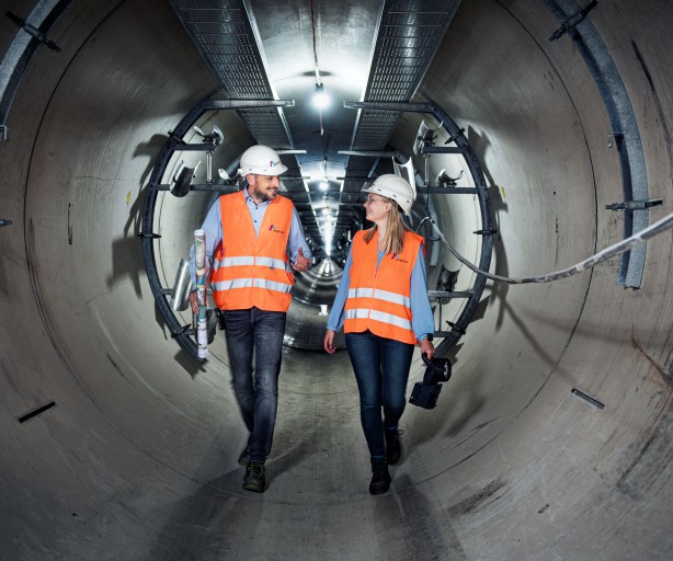 Das Foto zeigt eine Zentralperspektive im Inneren eines großen, runden Beton-Leitungstunnels.
Zentrale Motive: Zwei Personen, eine Frau und ein Mann, gehen nebeneinander durch den Tunnel auf den Betrachter zu. Beide tragen Schutzausrüstung bestehend aus weißen Schutzhelmen mit Amprion-Logo, leuchtend orangenen Warnwesten über blauen Hemden bzw. Blusen sowie dunkle Jeans.
Aktion und Details: Der Mann auf der linken Seite trägt eine zusammengerollte technische Zeichnung unter dem Arm und spricht mit seiner Kollegin. Die Frau rechts hält eine schwarze Handlampe. Beide lächeln sich im Gehen an.
Der Tunnel: Die Tunnelwände bestehen aus hellgrauen Betonsegmenten. An der Decke verläuft eine metallene Kabelpritsche, an der in regelmäßigen Abständen helle Arbeitsleuchten montiert sind. Diese erzeugen einen starken Tiefeneffekt, der den Blick in die scheinbar endlose Ferne des Tunnels lenkt. An den Seitenwänden sind metallene Halterungen für Kabel oder Rohrleitungen angebracht.
Atmosphäre: Die Szene wirkt professionell, kooperativ und unterstreicht die Dimensionen unterirdischer Infrastrukturprojekte.
