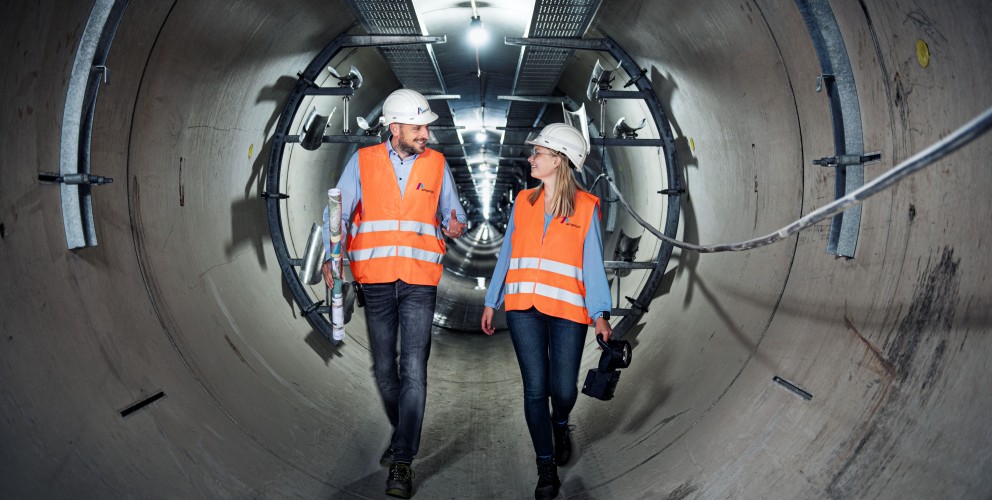 Das Foto zeigt eine Zentralperspektive im Inneren eines großen, runden Beton-Leitungstunnels.
Zentrale Motive: Zwei Personen, eine Frau und ein Mann, gehen nebeneinander durch den Tunnel auf den Betrachter zu. Beide tragen Schutzausrüstung bestehend aus weißen Schutzhelmen mit Amprion-Logo, leuchtend orangenen Warnwesten über blauen Hemden bzw. Blusen sowie dunkle Jeans.
Aktion und Details: Der Mann auf der linken Seite trägt eine zusammengerollte technische Zeichnung unter dem Arm und spricht mit seiner Kollegin. Die Frau rechts hält eine schwarze Handlampe. Beide lächeln sich im Gehen an.
Der Tunnel: Die Tunnelwände bestehen aus hellgrauen Betonsegmenten. An der Decke verläuft eine metallene Kabelpritsche, an der in regelmäßigen Abständen helle Arbeitsleuchten montiert sind. Diese erzeugen einen starken Tiefeneffekt, der den Blick in die scheinbar endlose Ferne des Tunnels lenkt. An den Seitenwänden sind metallene Halterungen für Kabel oder Rohrleitungen angebracht.
Atmosphäre: Die Szene wirkt professionell, kooperativ und unterstreicht die Dimensionen unterirdischer Infrastrukturprojekte.
