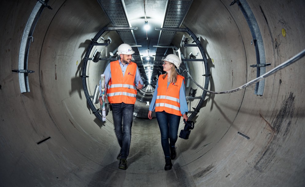 Das Foto zeigt eine Zentralperspektive im Inneren eines großen, runden Beton-Leitungstunnels.
Zentrale Motive: Zwei Personen, eine Frau und ein Mann, gehen nebeneinander durch den Tunnel auf den Betrachter zu. Beide tragen Schutzausrüstung bestehend aus weißen Schutzhelmen mit Amprion-Logo, leuchtend orangenen Warnwesten über blauen Hemden bzw. Blusen sowie dunkle Jeans.
Aktion und Details: Der Mann auf der linken Seite trägt eine zusammengerollte technische Zeichnung unter dem Arm und spricht mit seiner Kollegin. Die Frau rechts hält eine schwarze Handlampe. Beide lächeln sich im Gehen an.
Der Tunnel: Die Tunnelwände bestehen aus hellgrauen Betonsegmenten. An der Decke verläuft eine metallene Kabelpritsche, an der in regelmäßigen Abständen helle Arbeitsleuchten montiert sind. Diese erzeugen einen starken Tiefeneffekt, der den Blick in die scheinbar endlose Ferne des Tunnels lenkt. An den Seitenwänden sind metallene Halterungen für Kabel oder Rohrleitungen angebracht.
Atmosphäre: Die Szene wirkt professionell, kooperativ und unterstreicht die Dimensionen unterirdischer Infrastrukturprojekte.
