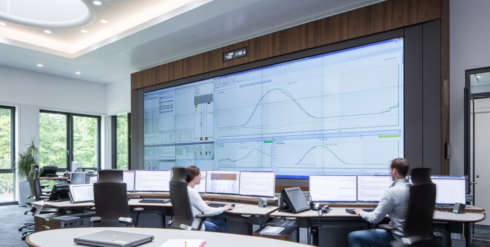 Zentrales Element: Eine massive, raumhohe Videowand dominiert den Hintergrund. Sie ist in eine dunkle Wand mit Holzoberkante eingefasst. Auf den Bildschirmen sind komplexe Datenvisualisierungen zu sehen, darunter mehrere große Graphen mit geschwungenen Kurven (Lastverläufe), Tabellen und technische Diagramme. Eine kleine Digitaluhr über der Wand zeigt die Zeit an.
Arbeitsplätze: Im Mittelgrund sitzen eine Frau und ein Mann mit dem Rücken zum Betrachter an einem bogenförmigen Schreibtischsystem. Vor ihnen stehen jeweils drei bis vier schmale Computermonitore. Sie sitzen auf ergonomischen, dunklen Bürostühlen und bedienen Tastaturen.
Raumgestaltung: Der Raum ist hell und sachlich gestaltet. Die Decke weist große, ovale Lichtelemente und integrierte Spotlights auf. Links bieten große Fensterfronten einen Blick auf grüne Bäume im Außenbereich. Im Vordergrund ist ein Teil eines ovalen Besprechungstisches mit einem geschlossenen Laptop zu sehen.
