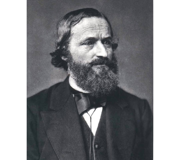 Das Bild zeigt ein historisches Porträt von Gustav Robert Kirchhoff (1824–1887), einem deutschen Physiker, der bedeutende Beiträge zur Elektrodynamik und Spektroskopie leistete. Kirchhoff hat ein ernstes Gesicht, trägt einen dichten, welligen Vollbart und kurzes, leicht gewelltes Haar. Er ist in einem dunklen Anzug mit einer Fliege und einem Hemd gekleidet, das den Stil des 19. Jahrhunderts widerspiegelt. Der Blick ist leicht nach rechts gerichtet. Das Schwarz-Weiß-Foto unterstreicht den zeitlichen Kontext und die historische Bedeutung der Aufnahme.