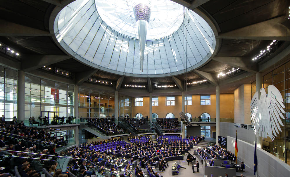 Panoramaaufnahme des voll besetzten Plenarsaals im Deutschen Bundestag während einer Sitzung, mit dem Bundesadler an der Wand im Vordergrund.