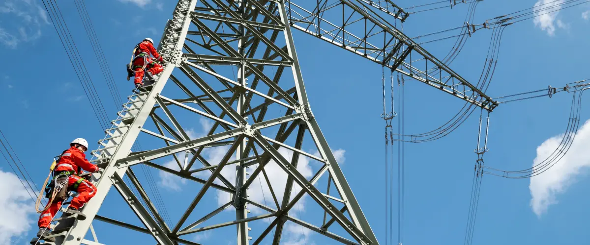 Zwei Menschen in roter Schutzkleidung und Helmen steigen an einem Hochspannungsmast senkrecht empor. Die massiven Stahlträger des Masts erstrecken sich diagonal ins Bild, darüber verlaufen mehrere Stromleitungen. Blauer Himmel mit wenigen weißen Wolken im Hintergrund.
