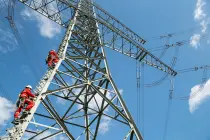 Zwei Menschen in roter Schutzkleidung und Helmen steigen an einem Hochspannungsmast senkrecht empor. Die massiven Stahlträger des Masts erstrecken sich diagonal ins Bild, darüber verlaufen mehrere Stromleitungen. Blauer Himmel mit wenigen weißen Wolken im Hintergrund.