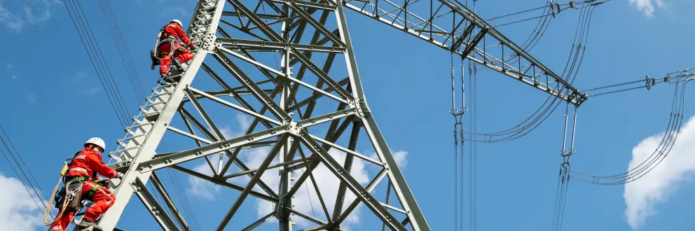 Zwei Menschen in roter Schutzkleidung und Helmen steigen an einem Hochspannungsmast senkrecht empor. Die massiven Stahlträger des Masts erstrecken sich diagonal ins Bild, darüber verlaufen mehrere Stromleitungen. Blauer Himmel mit wenigen weißen Wolken im Hintergrund.
