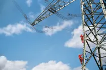 Zwei Menschen in roter Schutzkleidung und Helmen steigen an einem Hochspannungsmast senkrecht empor. Die massiven Stahlträger des Masts erstrecken sich diagonal ins Bild, darüber verlaufen mehrere Stromleitungen. Blauer Himmel mit wenigen weißen Wolken im Hintergrund.