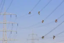 Mehrere fahrbare Arbeitskörbe mit Elektrikern hängen parallel an Stromleitungen zwischen mehreren Hochspannungsmasten. Die Körbe sind gleichmäßig verteilt und scheinen in der Luft zu schweben. Der Himmel ist klar blau.
