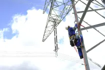 Ein Arbeiter in blauer Schutzkleidung klettert gesichert mit Seil und Karabiner an einem Hochspannungsmast empor. Oben im Bild sind Isolatoren und Leitungen zu erkennen. Der Himmel ist hellblau mit weißen Wolken.