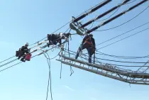Zwei Monteure in Sicherheitsausrüstung arbeiten direkt an Stromleitungen in luftiger Höhe. Sie stehen auf einem abgesicherten Steg, der an den Leitungen befestigt ist. Weitere Seile, Isolatoren und technische Vorrichtungen sind sichtbar. Blauer Himmel im Hintergrund.