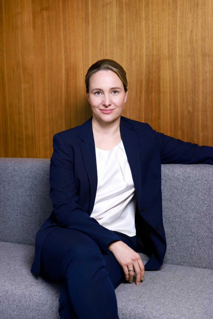 Katrin Hilmer, Mitglied der Amprion-Geschäftsführung: Sie sitzt auf einem grauen Sofa, trägt einen dunkelblauen Hosenanzug mit weißer Bluse und blickt freundlich in die Kamera. Im Hintergrund eine Holzvertäfelung.