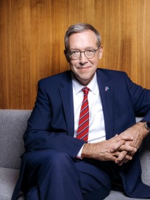 Ein Mann mittleren Alters mit kurzen grauen Haaren und Brille sitzt lächelnd vor einem holzvertäfeltem Hintergrund. Er trägt einen dunkelblauen Anzug, ein weißes Hemd und eine rote Krawatte mit feiner Struktur. Sein Ausdruck wirkt freundlich und professionell.