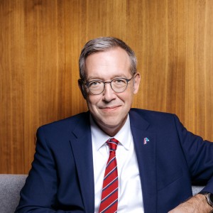 Ein Mann mittleren Alters mit kurzen grauen Haaren und Brille sitzt lächelnd vor einem holzvertäfeltem Hintergrund. Er trägt einen dunkelblauen Anzug, ein weißes Hemd und eine rote Krawatte mit feiner Struktur. Sein Ausdruck wirkt freundlich und professionell.