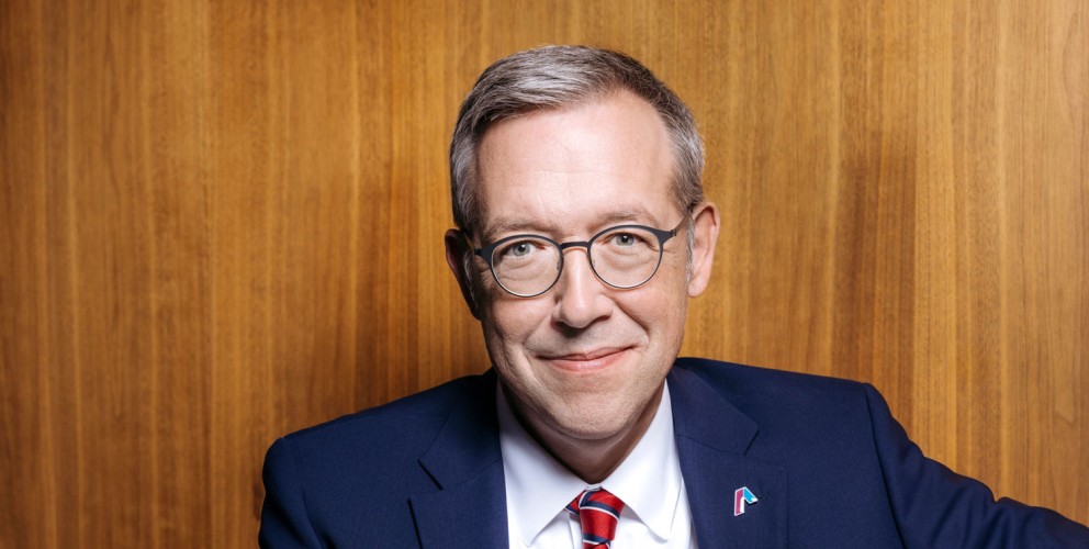 Ein Mann mittleren Alters mit kurzen grauen Haaren und Brille sitzt lächelnd vor einem holzvertäfeltem Hintergrund. Er trägt einen dunkelblauen Anzug, ein weißes Hemd und eine rote Krawatte mit feiner Struktur. Sein Ausdruck wirkt freundlich und professionell.