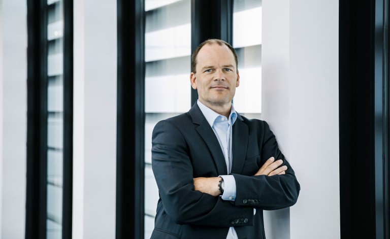 Ein Mann mittleren Alters mit kurzen Haaren steht mit verschränkten Armen in einem modernen Bürogebäude. Er trägt einen dunklen Anzug und ein hellblaues Hemd. Sein Gesichtsausdruck ist selbstbewusst und freundlich. Der Hintergrund zeigt große Fenster mit horizontalen Jalousien, durch die Tageslicht fällt, sowie weiße Wandflächen.