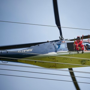 Das Foto aus der Froschperspektive zeigt einen unmittelbar neben der Leitung fliegenden, vierblättrigen Hubschrauber mit eingezogenem Fahrwerk. In der Tür des blauen Helikopters sitzt ein Monteur in rotem Overall, welche Vogelschutzmarkierungen anbringt.
