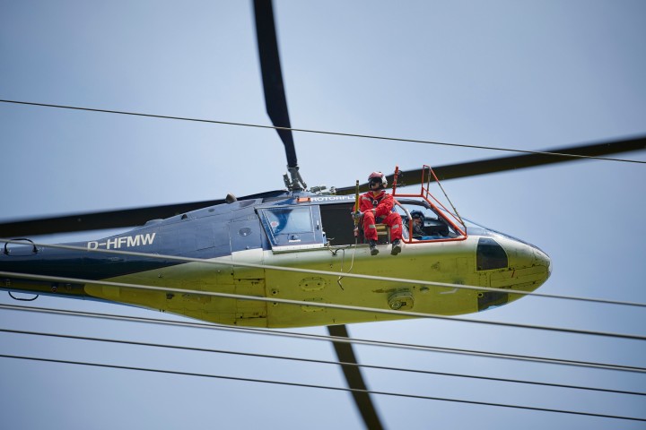 Das Foto aus der Froschperspektive zeigt einen unmittelbar neben der Leitung fliegenden, vierblättrigen Hubschrauber mit eingezogenem Fahrwerk. In der Tür des blauen Helikopters sitzt ein Monteur in rotem Overall, welche Vogelschutzmarkierungen anbringt.