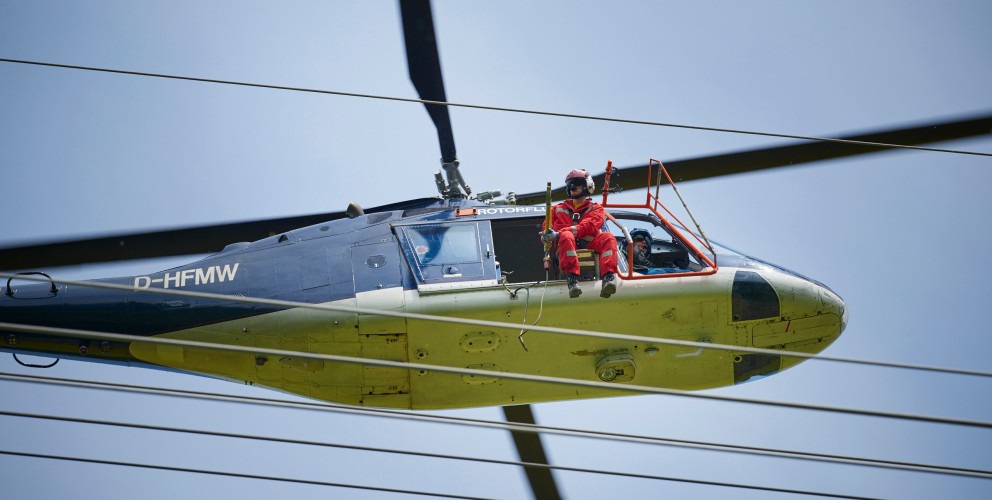 Das Foto aus der Froschperspektive zeigt einen unmittelbar neben der Leitung fliegenden, vierblättrigen Hubschrauber mit eingezogenem Fahrwerk. In der Tür des blauen Helikopters sitzt ein Monteur in rotem Overall, welche Vogelschutzmarkierungen anbringt.