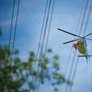 Das Foto aus der Froschperspektive zeigt einen unmittelbar neben der Leitung fliegenden, vierblättrigen Hubschrauber mit eingezogenem Fahrwerk. In der Tür des blauen Helikopters sitzt ein Monteur in rotem Overall, welche Vogelschutzmarkierungen anbringt.