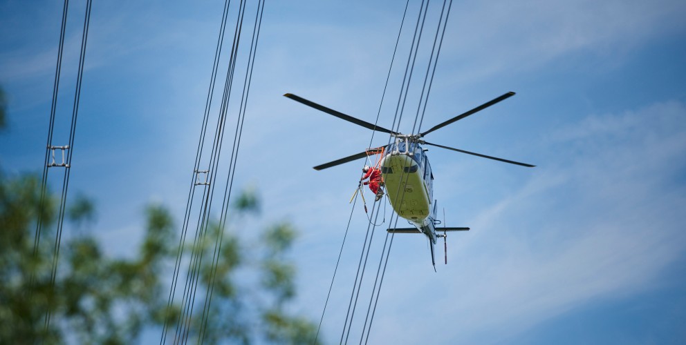 Das Foto aus der Froschperspektive zeigt einen unmittelbar neben der Leitung fliegenden, vierblättrigen Hubschrauber mit eingezogenem Fahrwerk. In der Tür des blauen Helikopters sitzt ein Monteur in rotem Overall, welche Vogelschutzmarkierungen anbringt.