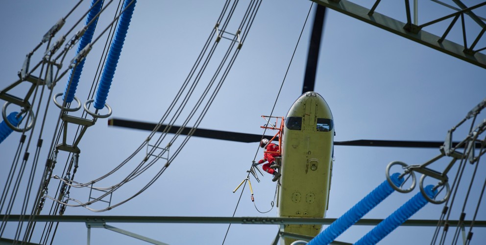 Das Foto aus der Froschperspektive zeigt einen unmittelbar neben der Leitung fliegenden, vierblättrigen Hubschrauber mit eingezogenem Fahrwerk. In der Tür des blauen Helikopters sitzt ein Monteur in rotem Overall, welche Vogelschutzmarkierungen anbringt.