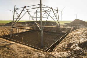 Baustelle für das Fundament eines neuen Hochspannungsmasts in ländlicher Umgebung. Sichtbar ist ein großer rechteckiger Aushub mit Bewehrungsstahl für das Betonfundament, über dem ein Teil des Stahlgerüsts des Masts bereits montiert ist. Rund um die Baustelle stehen Absperrgitter. Im Hintergrund sind grüne Felder, Erdhügel und mehrere Windkraftanlagen zu sehen, was auf eine Verbindung zu erneuerbarer Energie hinweisen könnte. Die Lichtstimmung ist warm, vermutlich am Morgen oder späten Nachmittag.
