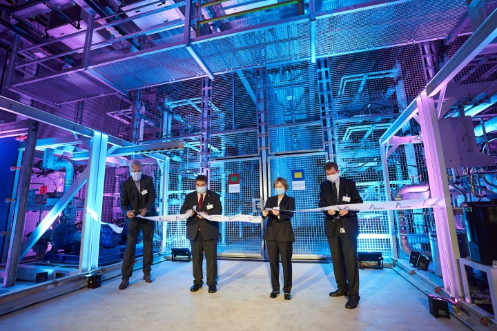 Vier Personen in Businesskleidung stehen in einer hochmodernen, industriellen Anlage mit futuristischer blauer und violetter Beleuchtung. Sie tragen Schutzmasken und schneiden gemeinsam ein weißes Band mit Firmenlogo durch – eine klassische Zeremonie zur Eröffnung einer neuen Einrichtung oder technischen Anlage. Hinter ihnen sind Gitterstrukturen, Rohre und technische Installationen sichtbar. Die Szenerie wirkt technisch-innovativ, und die Veranstaltung scheint einen feierlichen Anlass zu markieren.