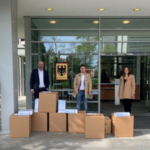 Fünf Personen stehen vor einem modernen Gebäude mit Glasfassade, über dessen Eingang das Schild der Bundesnetzagentur mit dem Bundesadler hängt. Sie sind geschäftlich gekleidet, einige tragen Anzüge, andere Mäntel. Vor ihnen sind mehrere große Kartons aufgestapelt, aus denen Aktenordner und Dokumente herausragen. Zwei der Männer halten ebenfalls Unterlagen in der Hand. Die Gruppe wirkt offiziell und posiert für das Foto, vermutlich im Zusammenhang mit der Übergabe von Dokumenten an die Behörde.