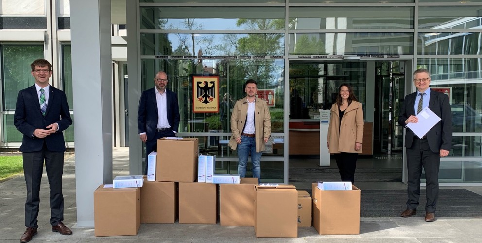 Fünf Personen stehen vor einem modernen Gebäude mit Glasfassade, über dessen Eingang das Schild der Bundesnetzagentur mit dem Bundesadler hängt. Sie sind geschäftlich gekleidet, einige tragen Anzüge, andere Mäntel. Vor ihnen sind mehrere große Kartons aufgestapelt, aus denen Aktenordner und Dokumente herausragen. Zwei der Männer halten ebenfalls Unterlagen in der Hand. Die Gruppe wirkt offiziell und posiert für das Foto, vermutlich im Zusammenhang mit der Übergabe von Dokumenten an die Behörde.