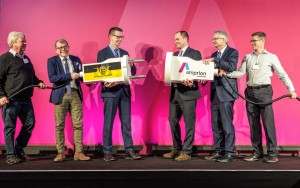 Das Foto zeigt sechs Männer, die auf einer Bühne vor einem leuchtend pinkfarbenen Hintergrund stehen. Sie sind in zwei Gruppen aufgeteilt und halten jeweils ein großes, weißes, stilisiertes Stromkabel mit einem überdimensionierten Stecker und einer ebensolchen Kupplung.