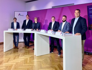 Das Bild zeigt sechs Personen, die hinter zwei modernen, weißen Tischen mit Namensschildern stehen. Sie posieren für ein Gruppenfoto in einem Veranstaltungsraum mit einem violetten Hintergrund, auf dem ein Logo und Text zu sehen sind. Alle tragen geschäftliche Kleidung, hauptsächlich Anzüge und Blazer. Die Atmosphäre wirkt professionell und freundlich, da alle lächeln. Der Raum hat einen hellen Holzfußboden, und das Setting erinnert an eine Podiumsdiskussion oder eine Vertragsunterzeichnung.