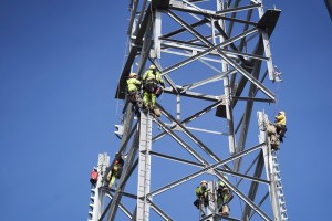 Mehrere Arbeiter in leuchtend gelben Schutzanzügen und Helmen klettern an einem hohen Stahlgittermast. Sie sind mit Klettergurten gesichert und benutzen Leitern sowie Seile, während sie an der Konstruktion arbeiten. Der Hintergrund zeigt einen strahlend blauen Himmel, was die Höhe des Strommasts besonders betont.