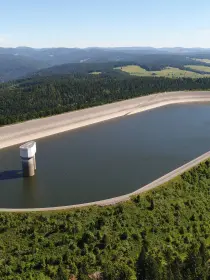 Ein großes, ovales Speicherbecken auf einem Hügel, umgeben von grüner Landschaft. Die Wasseroberfläche ist ruhig, und ein rundes Einlassbauwerk ragt aus dem Wasser. Die Wände des Beckens sind betoniert, und ein Weg führt entlang des Randes. Im Hintergrund sind bewaldete Hügel und offene Felder zu sehen.