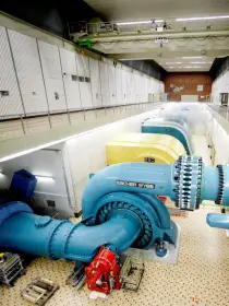 Eine langgestreckte Industriehalle mit mehreren großen, blauen Wasserturbinen von „Escher Wyss“. Die Turbinen sind mit gelben Generatoren verbunden. Ein Laufsteg mit Geländer verläuft entlang der Hallenwand. Die Decke ist mit zahlreichen Lichtern ausgestattet.