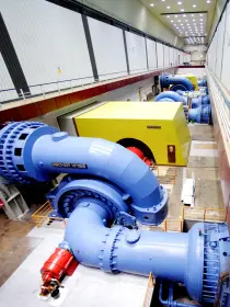 Ein weiteres Bild einer Maschinenhalle mit großen Wasserturbinen. Im Vordergrund ist eine blaue Turbine mit dem Schriftzug „Escher Wyss“, verbunden mit einem dicken Rohr. Dahinter sind weitere Maschinen, gelbe Generatoren und Steuerungselemente zu sehen. Die Halle ist lang und gut beleuchtet.