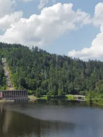 Eine Außenaufnahme eines Wasserkraftwerks an einem See. Im Hintergrund erhebt sich ein bewaldeter Hügel mit einer langen, steilen Rohrleitung, die von einem Gebäude auf der Hügelkuppe hinabführt. Das Kraftwerk befindet sich am Ufer des Sees und besteht aus 